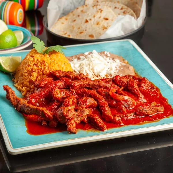 Red sauced meat strips with rice, beans, and lime on a blue plate, with tortillas in background.
