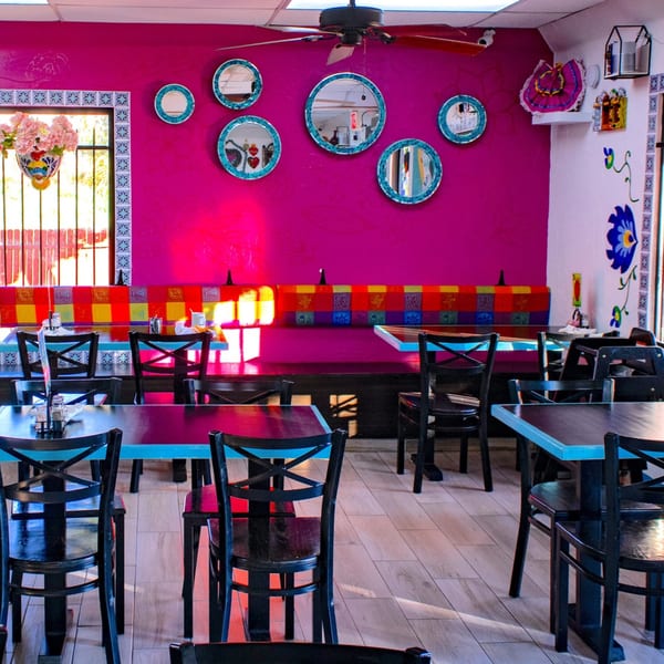 Vibrant restaurant interior with fuchsia walls, colorful banquettes, black tables, and decorative mirrors.