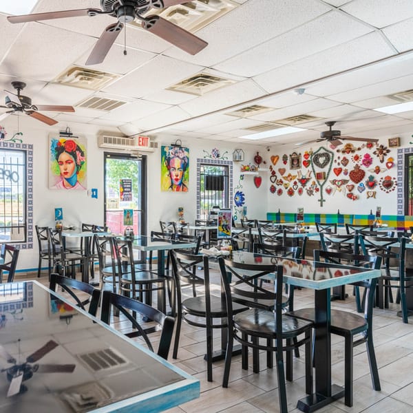 Brightly lit restaurant interior with black tables and chairs, colorful wall art, and ceiling fans.