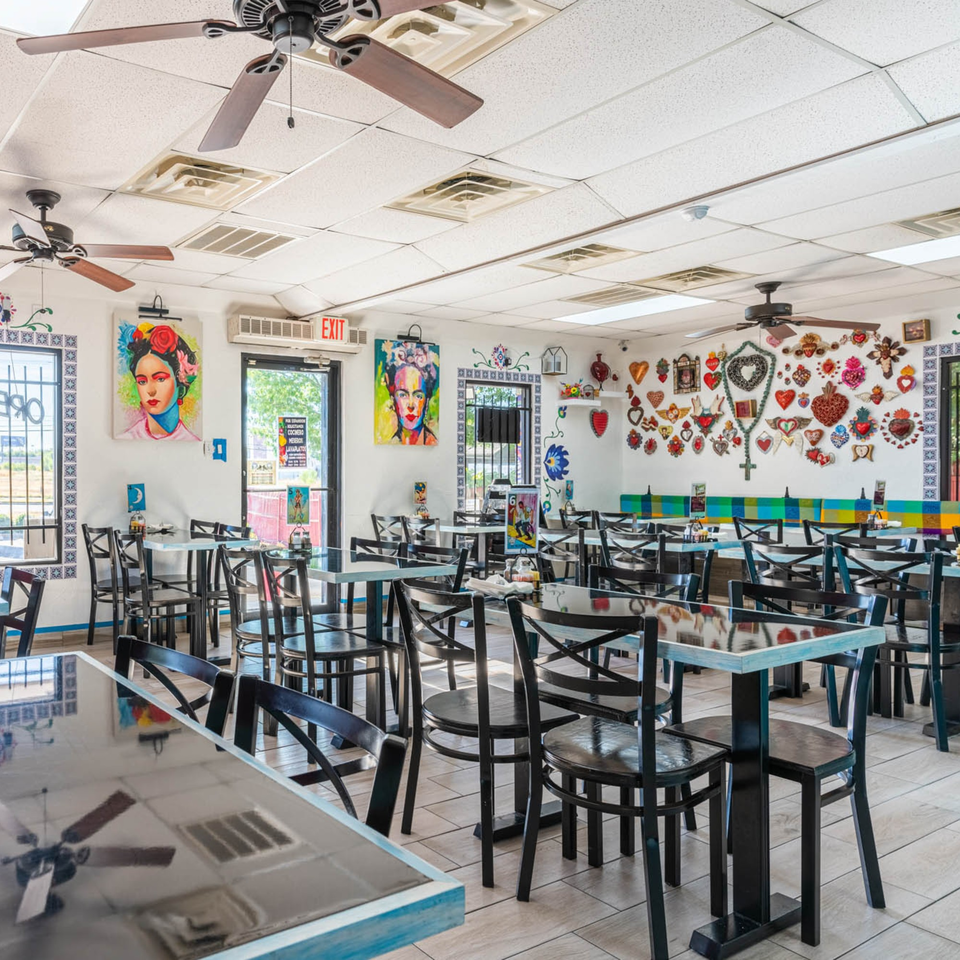 Brightly lit restaurant interior with black tables and chairs, colorful wall art, and ceiling fans.