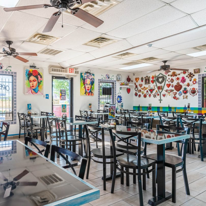 Brightly lit restaurant interior with black tables and chairs, colorful wall art, and ceiling fans.