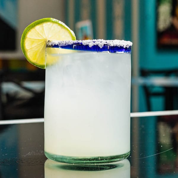 A margarita in a blue-rimmed glass with a lime wedge and salted rim on a dark table.