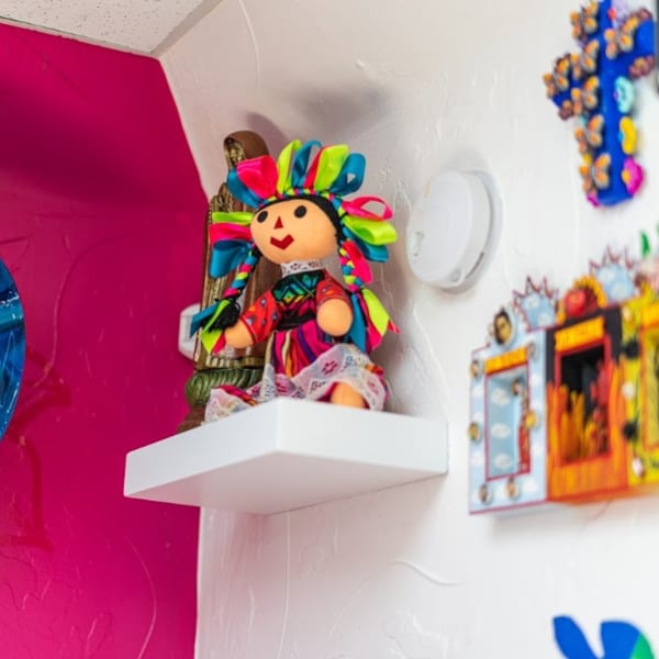 A colorful Mexican rag doll sits on a white shelf next to a religious statue and vibrant wall decor.