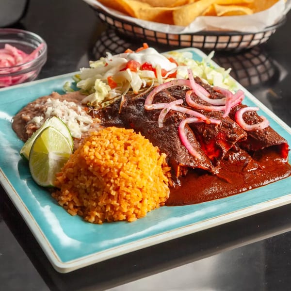 Three enchiladas covered in dark red sauce and pickled onions, with rice, beans, and lime wedges.