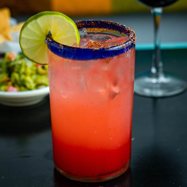 A vibrant red drink with ice, a lime wedge, and a chili-salted rim in a blue-rimmed glass.