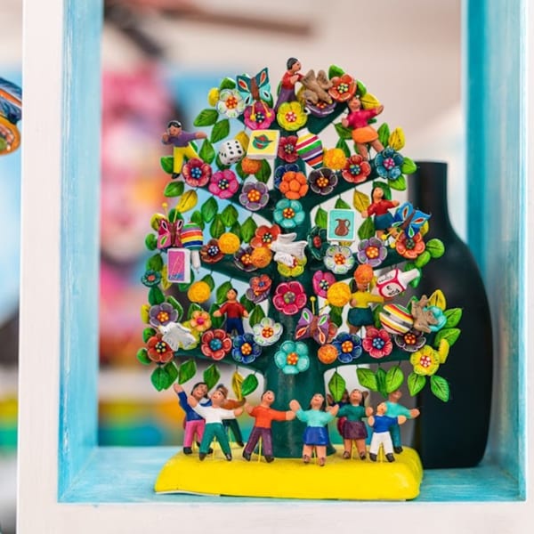 A colorful Tree of Life sculpture with miniature figures, flowers, and objects, displayed in a blue shelf.