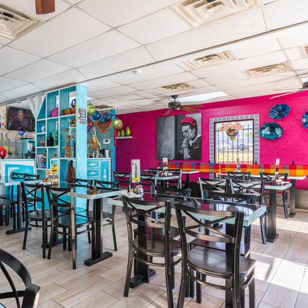 Vibrant Mexican restaurant interior with fuchsia walls, blue shelving, black tables, and a large Frida Kahlo portrait.