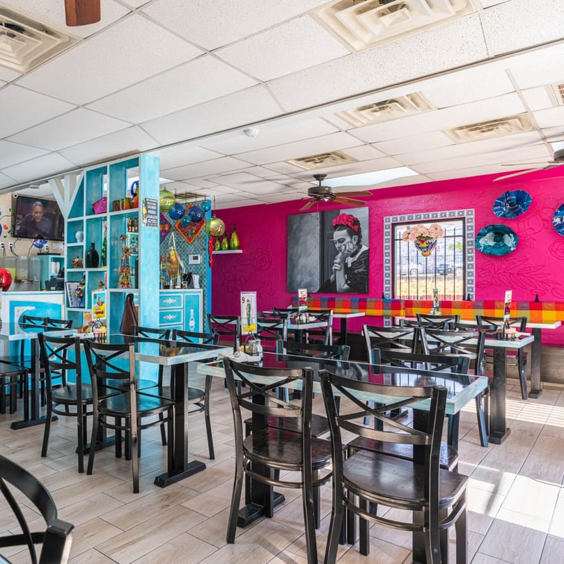 Vibrant Mexican restaurant interior with fuchsia walls, blue shelving, black tables, and a large Frida Kahlo portrait.